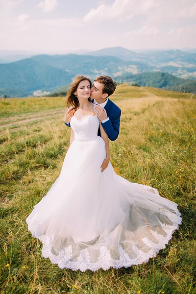 Joven pareja hermosa boda posando en el campo de hierba soleada con colinas forestales distantes y el cielo maravilloso como fondo —  Fotos de Stock