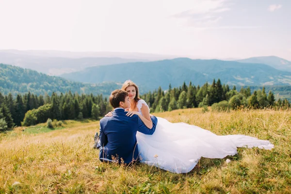 Novia recién casada en las manos de su nuevo marido sentado en la hierba amarilla en el campo soleado con majestuosas colinas forestales como fondo —  Fotos de Stock