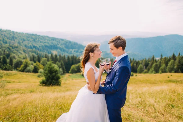 Joyeux jeunes mariés buvant du vin à l'extérieur, célébrant leur mariage. Superbes collines forestières comme arrière-plan — Photo