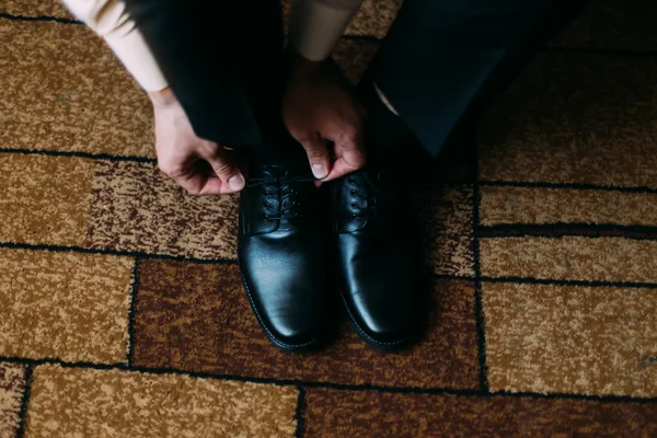 Close up de perna de homem e mãos amarrando laços de sapato preto elegante em pé no tapete com ornamento retangular — Fotografia de Stock