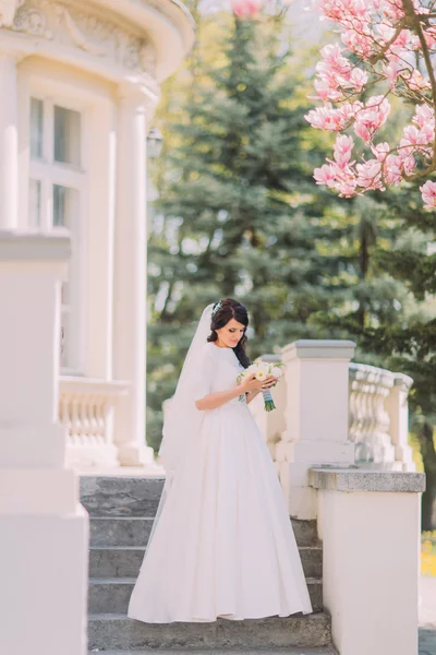 Noiva com buquê de casamento em pé no vintage edifício branco escadas traseiras sob ramo de magnólia florescente — Fotografia de Stock