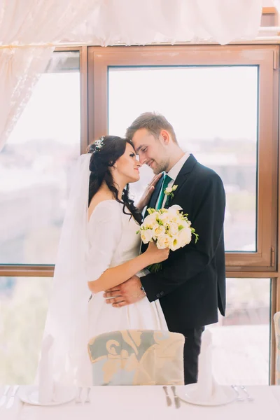 Bonito par elegante de recém-casados abraçando uns aos outros no interior do restaurante de luxo. Janela brilhante como fundo — Fotografia de Stock