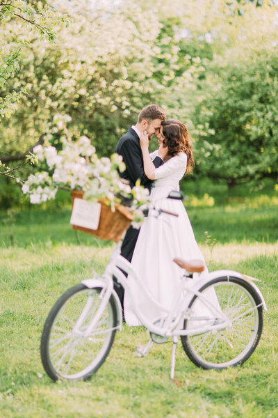Sensual young newlywed couple holding each other in park. Bicycle with wedding decoration on foreground