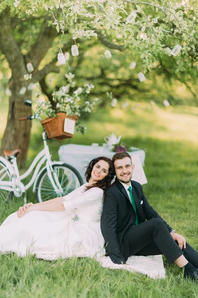 Счастливая невеста и жених сидят на траве спина к спине в парке с велосипедом рядом с деревом на заднем плане — стоковое фото