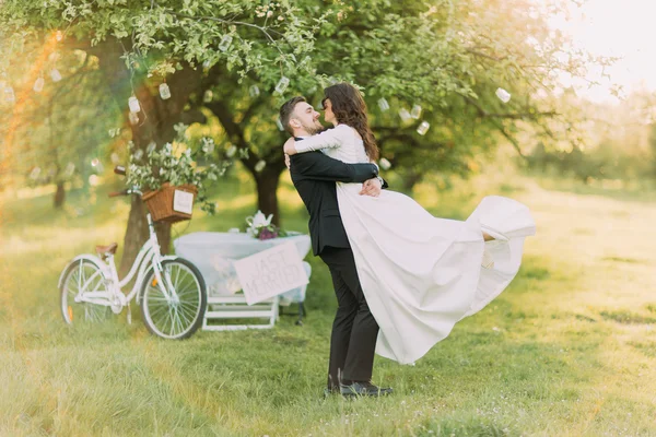 Feliz pareja de recién casados bailando en el césped en el verde parque soleado. Bicicleta cerca del árbol decorado al fondo —  Fotos de Stock