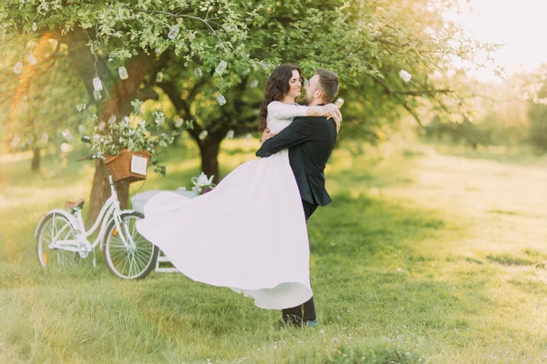 Happy acabou de se casar dançando no gramado no parque ensolarado verde. Bicicleta perto de árvore decorada no fundo — Fotografia de Stock