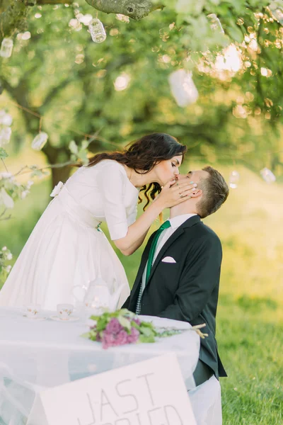 Romantická okurková v parku. Hravý nevěsta líbá svého milího nového manžela, zatímco sedí u stolu — Stock fotografie