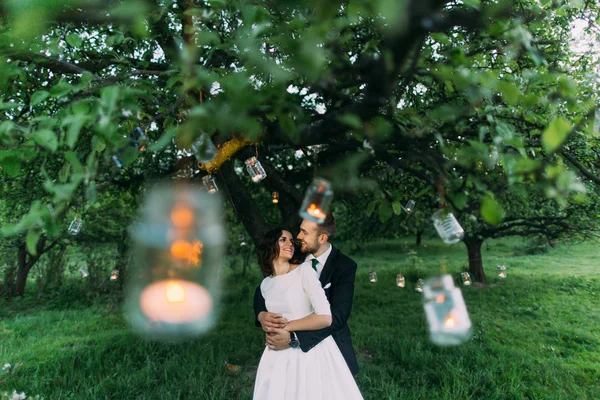 Bela noiva e seu noivo amoroso no parque da noite segurando um ao outro sob a árvore decorada com muitas lanternas — Fotografia de Stock