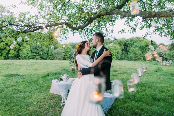 Beautiful bride and groom in evening park embracing under tree decorated with many lanterns — Stock Photo, Image