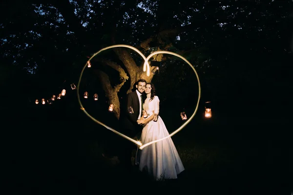 Hermosos novios en el parque nocturno sosteniéndose entre sí bajo el árbol decorado con muchas linternas. Corazón pintado de luz —  Fotos de Stock