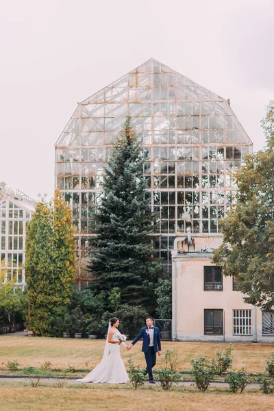 Belle mariée avec robe de mariée blanche et marié en costume bleu élégant marchant en plein air dans un parc. Grande serre est à l'arrière-plan — Photo