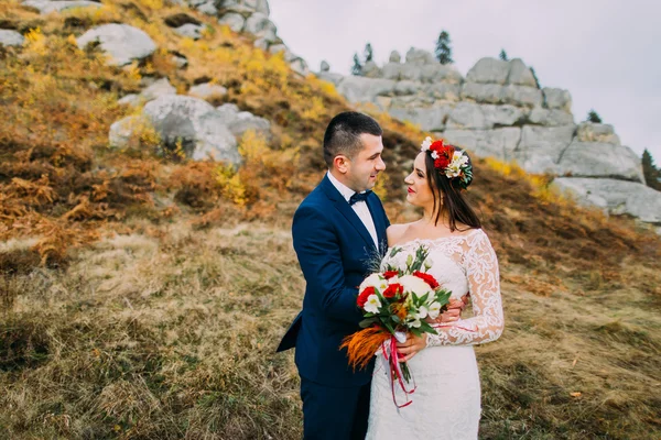 Mooie bruidspaar op idyllische pastoraal landschap met rotsen en hek als backround. Bruidegom in stijlvolle blauwe pak, witte gekleed bruid holding boeket rozen — Stockfoto