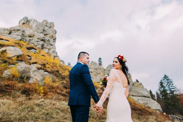 Bello sposo in elegante abito blu che si tiene per mano con sposa vestita di bianco su idilliaco paesaggio pastorale. Sullo sfondo rocce maestose — Foto Stock