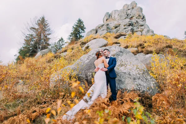 Şık mavi yakışıklı damat takım elbise kucaklayan beyaz giyimli gelin gül buketi artalanları olarak büyük kayalar ile pastoral açık peyzaj üzerinde tutarak. Düşük açılı görünüş