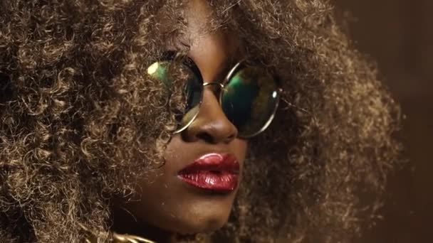 Primer plano del modelo femenino afroamericano dorado mágico en gafas de sol masivas con maquillaje brillante brillante, peinado dorado brillante y grandes labios rojos posando en el fondo del estudio — Vídeos de Stock