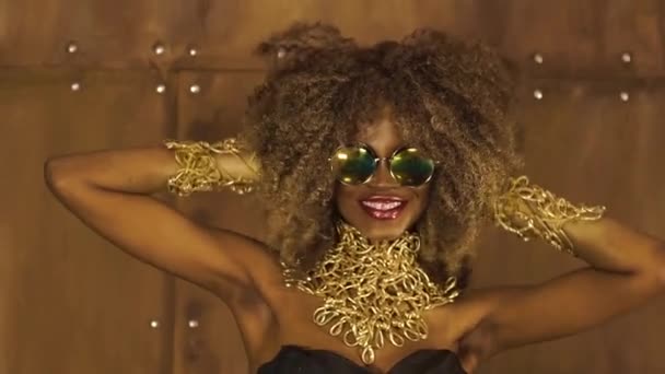 Modelo femenino afroamericano surrealista mágico dorado en gafas de sol masivas con maquillaje brillante brillo, peinado dorado brillante y grandes labios rojos posando y riendo en el fondo del estudio — Vídeos de Stock