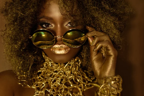 Close-up de magia dourada afro-americana modelo feminino em óculos de sol maciços com maquiagem brilhante brilho, penteado brilhante e grandes lábios vermelhos posando no fundo do estúdio Fotos De Bancos De Imagens