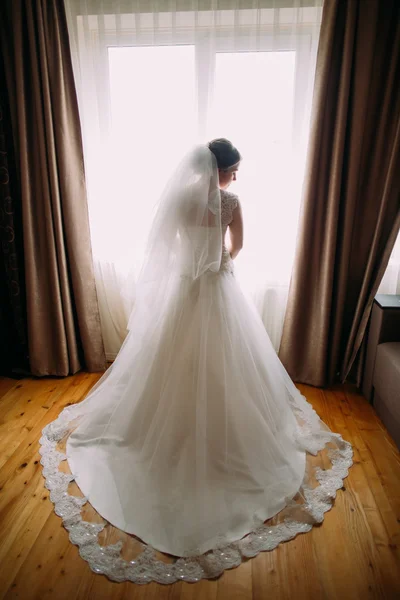 Belle mariée avec un long voile debout contre les lumières de fenêtre — Photo