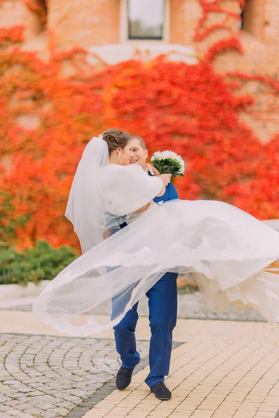 Young husband holding beautiful wife on his hands. Red creeping plant as background — Stock Photo, Image