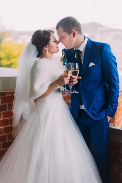 Recém-casados comemorando seu casamento bebendo champanhe e indo para beijar de pé perto da parede de tijolo — Fotografia de Stock