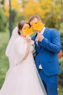Yeni evli çift açık havada poz. Gençler yüzlerini sonbahar yaprakları arkasında saklanıyor