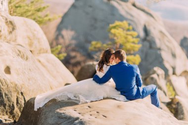 Romantik yeni evli çift, güneş ışığında üzerinde kayalık uçurum oturan kaya yayılan gelinler uzun elbise
