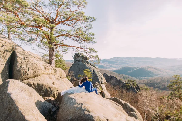 Casal recém-casado romântico sentado em penhasco com paisagem de montanha. Vista de pares para trás — Fotografia de Stock