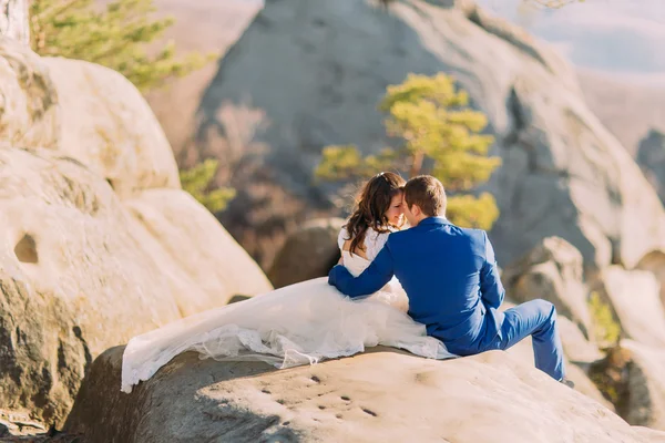 Romántica pareja de recién casados sentados a la luz del sol en el acantilado rocoso, novias vestido largo que extiende la roca —  Fotos de Stock