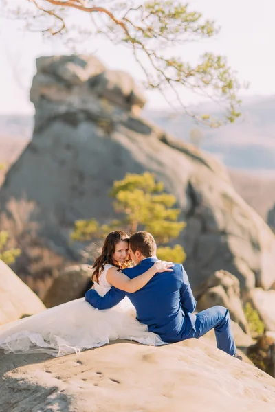 Romantik yeni evli çift, güneş ışığında üzerinde kayalık uçurum oturan kaya yayılan gelinler uzun elbise — Stok fotoğraf