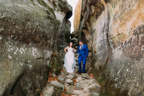Junge romantische Braut mit ihrem liebenden Bräutigam posiert auf Bergpfad in verwitterter Felsspalte — Stockfoto