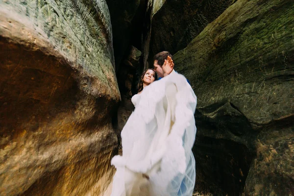 Young and romantic bride in white wedding dress with her loving groom posing at darkened rock cleft — Stock Photo, Image