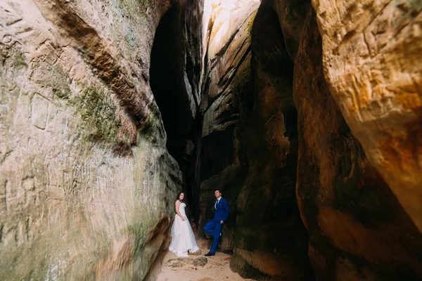 Jeune mariée romantique avec une longue robe blanche et son marié posant à la fente rocheuse sombre — Photo