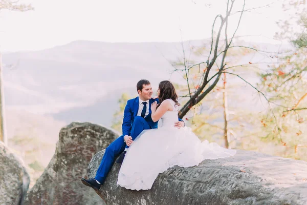 Romántica pareja recién casada sentada en un acantilado rocoso, novias vestido largo esparciendo roca. Colinas paisaje —  Fotos de Stock