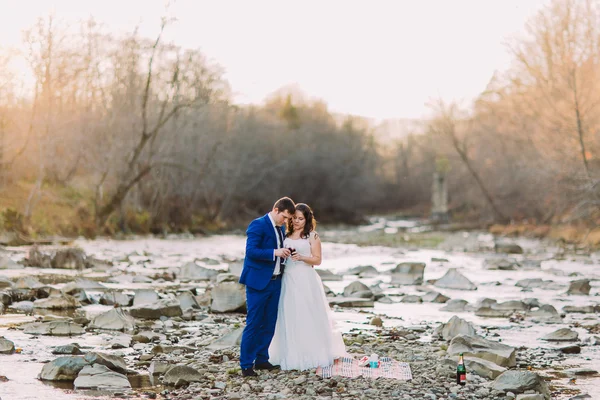 Romantické mladé novomanžele pití vína na břehu řeky kamenité, oblázkové s forest hills a stream — Stock fotografie