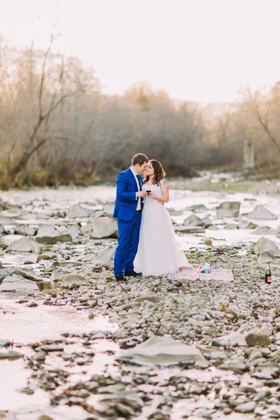 Romantica giovane coppia di sposi che beve vino sulla riva rocciosa del fiume di ghiaia con colline boschive e ruscello — Foto Stock
