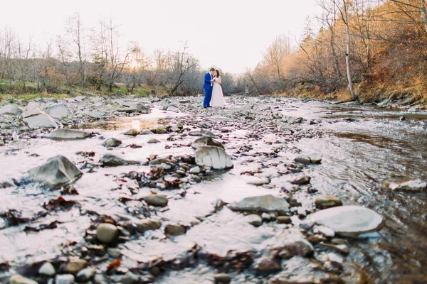 Romántica pareja de novias jóvenes bebiendo vino en la orilla rocosa del río guijarro con colinas forestales y arroyo — Foto de Stock