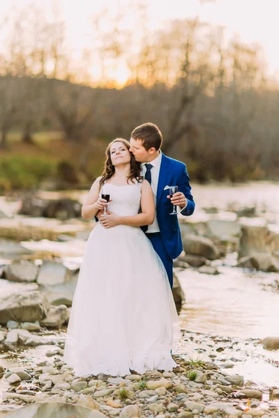 Romántica pareja de recién casados bebiendo vino tinto en la orilla del río guijarro con colinas forestales y arroyo — Foto de Stock