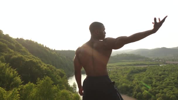 Vista posteriore. Muscoloso uomo atletico afroamericano con busto nudo in posa sulla roccia e mostrando i muscoli. Verde montagna natura sfondo — Video Stock