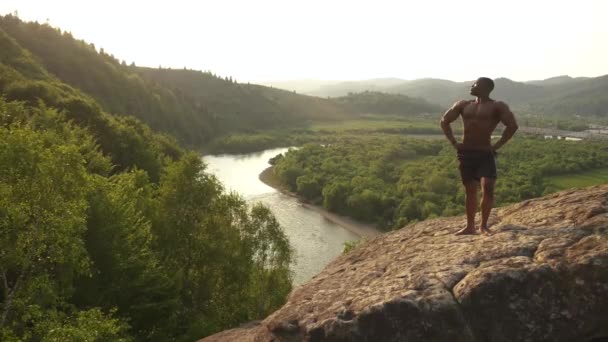 Der afrikanisch-amerikanische Bodybuilder steht auf einem Felsen vor dem blauen wolkenverhangenen Himmel und genießt die Aussicht. Moment der Harmonie mit der Natur — Stockvideo