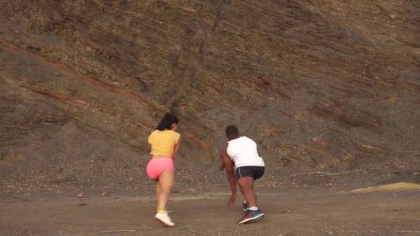 Молода змішана пара робить вправи на відкритому повітрі на кам'яному пляжі. Сексуальна брюнетка розтягується з особистим тренером з фітнесу чорношкірого афроамериканця. Скелястий гірський фон — стокове відео
