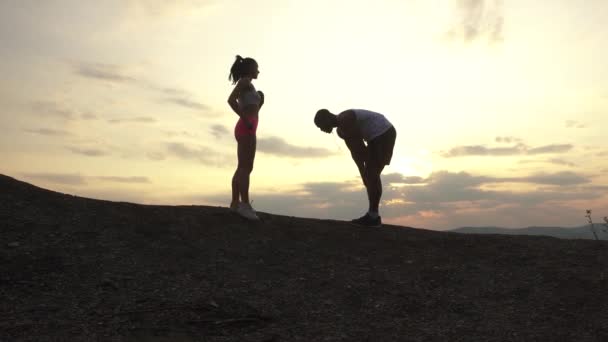Silhouetten van gemengd ras paar fitness-instructeurs doen oefeningen buiten op de achtergrond van de rocky mountains op zonsondergang. Sport fitness concept — Stockvideo