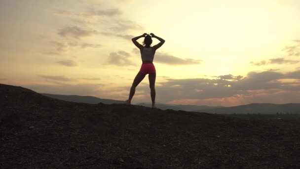 Вид ззаду сексуальна м'язиста жінка-інструктор з ідеальною дупою, що тягнеться на відкритому повітрі під час заходу сонця на вершині гори. Момент гармонії з природою — стокове відео