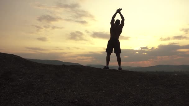 Silhouet van gespierde bodybuilder met perfect lichaam poseren bij de zonsopgang of zonsondergang in bergen. Knappe sterke man toont zijn spieren op achteraanzicht — Stockvideo