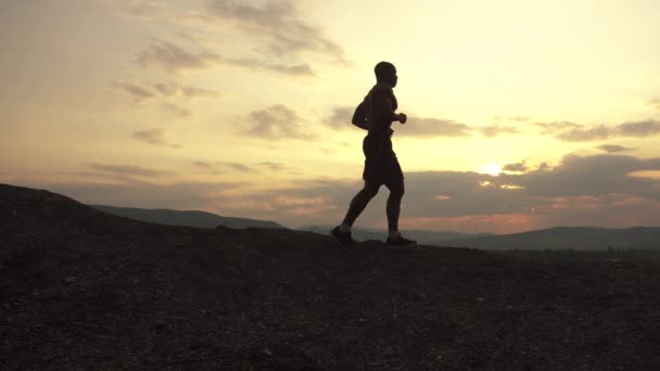 Sidovy på silhuett av afrikansk amerikansk atlet jogging på solnedgången på berget toppen. Molnigt Sky bakgrund, utbildning utomhus koncept — Stockvideo