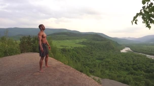 Widok z boku afrykańskiego amerykańskiego budowniczego ciała z doskonałym ciałem stwarzających na szczycie góry podczas porannego treningu na świeżym powietrzu. Wschód Słońca Niebo tle. Sport zdrowa koncepcja — Wideo stockowe