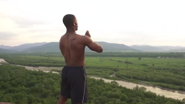 Muskularny African amerykański budowniczy ciała z modlitwą ręce rozciągające się na szczyt górski podczas porannego treningu na świeżym powietrzu. Wschód Słońca Niebo tle. Moment harmonii i szczęścia — Wideo stockowe