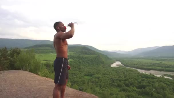 Vista lateral do homem musculoso americano africano sexy bebendo água após o treinamento esportivo. Paisagem de montanha verde deslumbrante no fundo — Vídeo de Stock