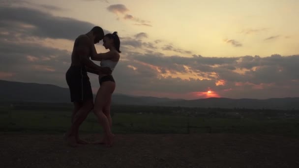 Silhouetten athletischer muskulöser Paare von Fitnesstrainern, die bei Sonnenuntergang auf dem Berggipfel tanzen. schönen bewölkten Himmel Hintergrund. Atemberaubender Moment — Stockvideo