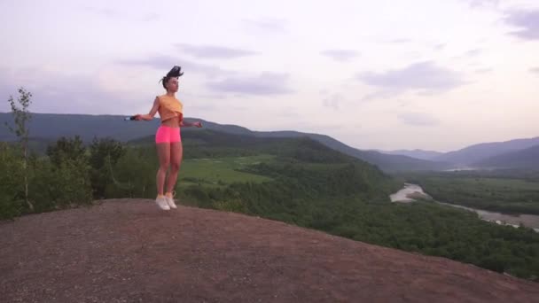 Joven morena activa saltando con una cuerda en la cima de la montaña. Fondo ladscape montaña verde — Vídeos de Stock