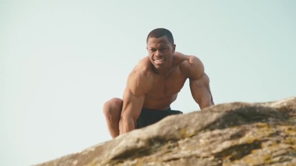 Bodybuilder noir noir émotionnel homme afro-américain avec torse nu posant sur la roche avec fond bleu ciel. Style nature sauvage — Video